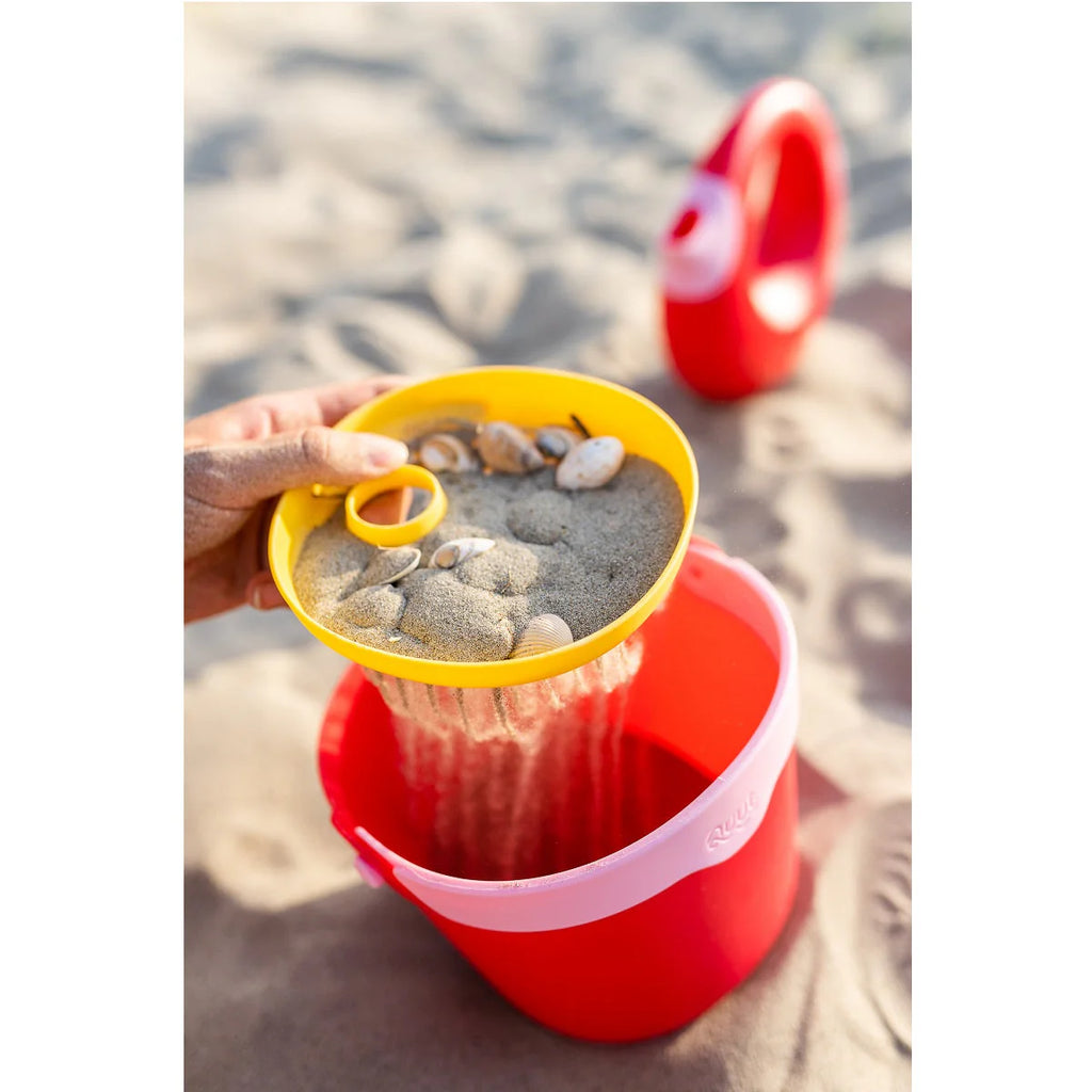 Red and yellow sand pail with sand and stones on a sandy beach