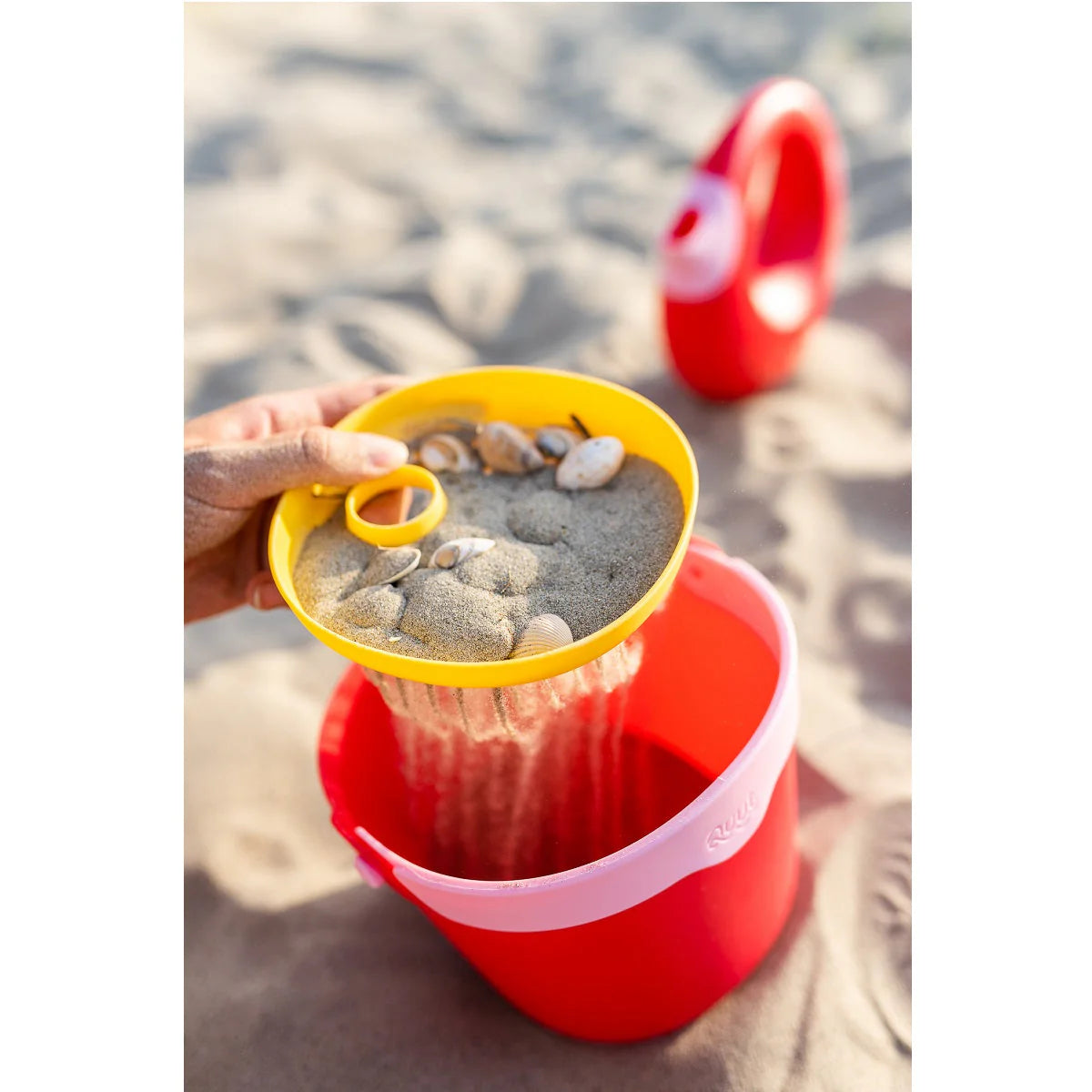 Red and yellow sand pail with sand and stones on a sandy beach